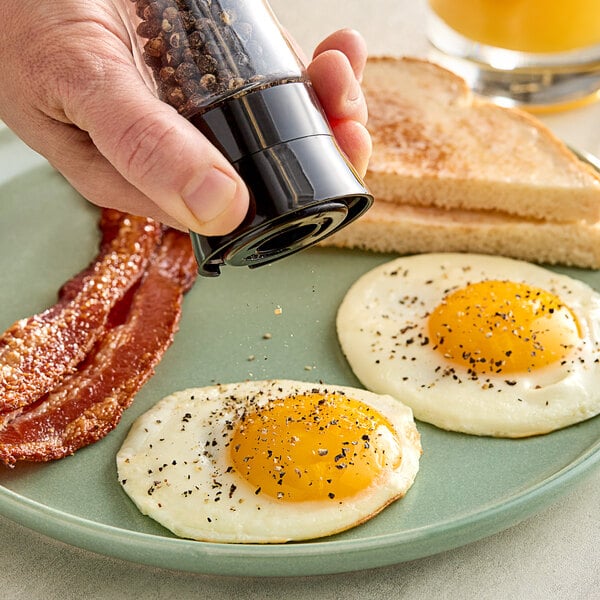 A hand using a Hometown Provisions pepper mill to sprinkle pepper on a plate of eggs, bacon, and toast.