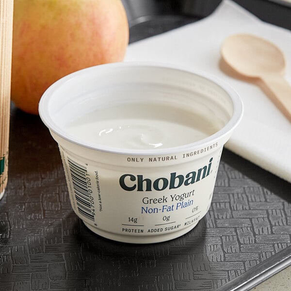 A white Chobani Non-Fat Plain Greek Yogurt container on a table with a spoon and apple.