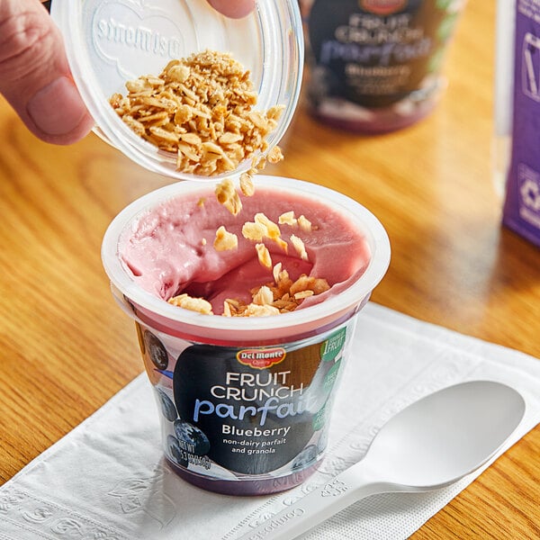 A hand pouring granola into a Del Monte Blueberry Fruit Crunch non-dairy yogurt cup.