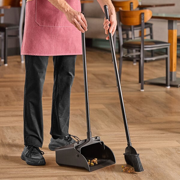 A person using an Advantage Supplies open-lid lobby dustpan and broom to sweep the floor.