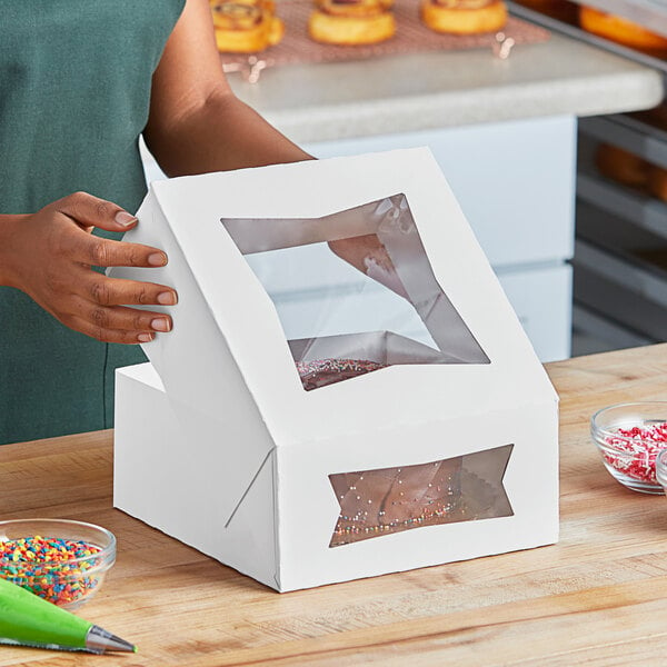 A woman using a green tip to decorate a cake with red and white sprinkles using a Southern Champion white bakery box with a window.