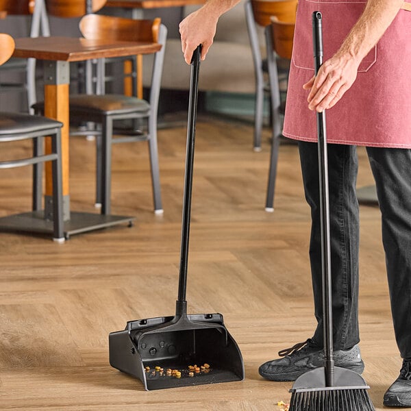 A person using an Advantage Supplies black open-lid dustpan to sweep a wood floor.