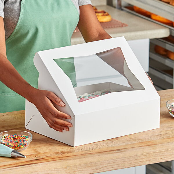 A person holding a white bakery box with a clear window displaying frosted pastries.