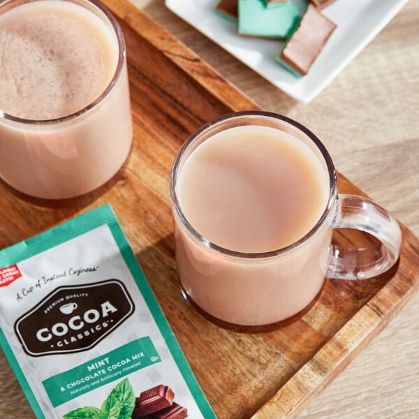 A glass mug of pink mint hot chocolate on a wooden tray with a package of Cocoa Classics Mint and Chocolate Cocoa mix.