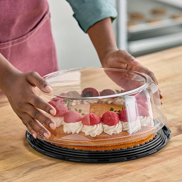 A person holding a cake in a D&W Fine Pack plastic cake display container with strawberries and whipped cream on a table in a bakery.