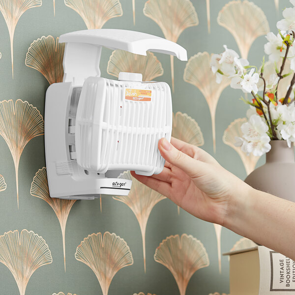 A hand replacing a white Oxy-Gen air freshener cartridge in a wall-mounted dispenser.