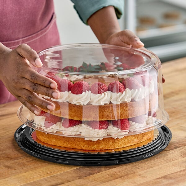 A person holding a cake with a clear plastic scalloped dome lid with strawberries and whipped cream on top.