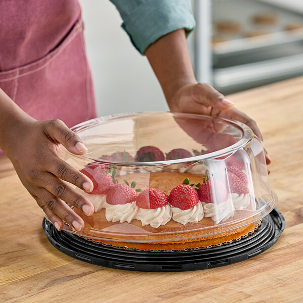 A person holding a D&W Fine Pack cake in a clear plastic container with a scalloped dome lid filled with strawberries and whipped cream.