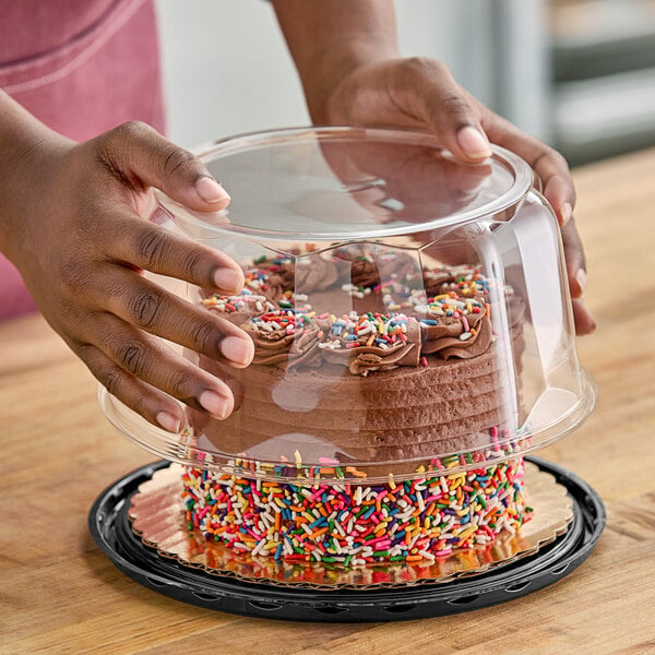 A person holding a frosted cake in a D&W Fine Pack clear plastic container with a clear scalloped dome lid.