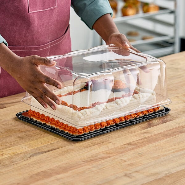 A person holding a D&W Fine Pack plastic container with a cake inside on a bakery display counter.