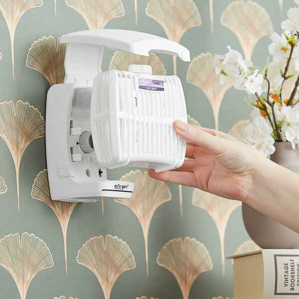 A hand replacing a white Oxy-Gen air freshener cartridge in a wall-mounted dispenser.