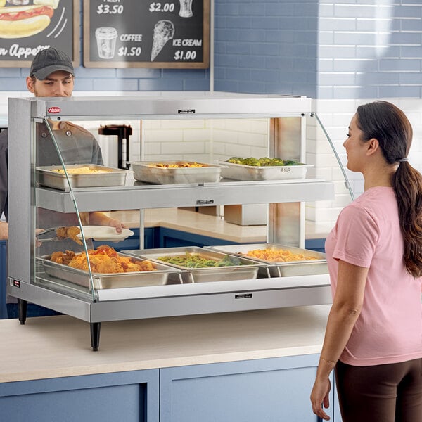 A woman in a pink shirt standing in front of a Hatco heated display case full of food.