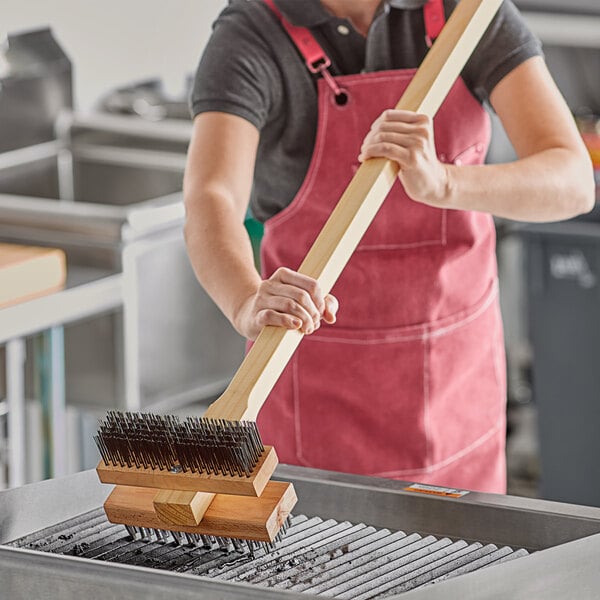 A woman in a red apron using a Choice double head carbon steel bristle grill brush to clean a grill.