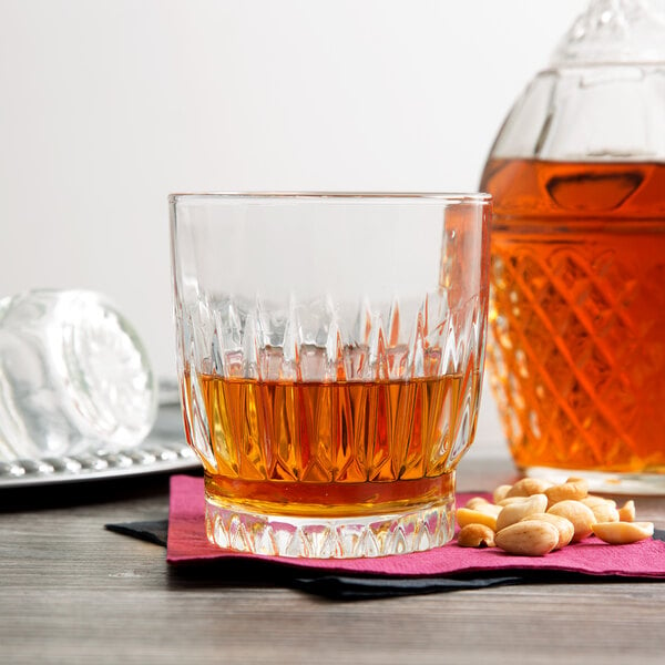 A Libbey Winchester rocks glass filled with amber liquid on a table next to a bottle of cashew nuts.