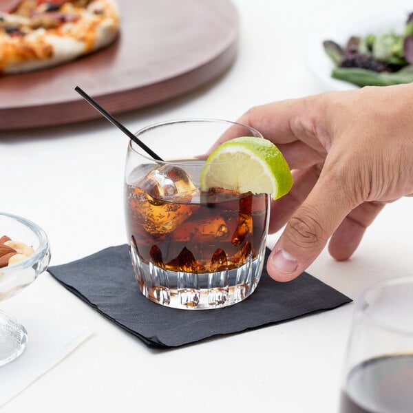 A hand holding a Reserve by Libbey Chisel Rocks glass with brown liquid and a lime wedge.