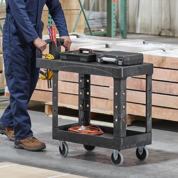 A man in a blue jumpsuit pushing a black Rubbermaid utility cart with tools on it.