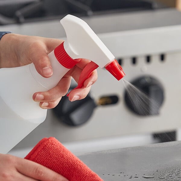 A person holding a Lavex red plastic spray bottle and spraying a professional kitchen counter.