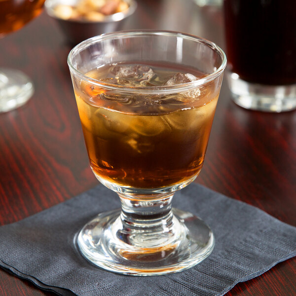 A Libbey footed rocks glass filled with a brown drink and ice on a napkin on a table.