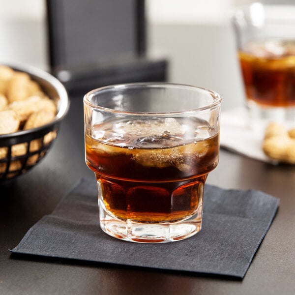 A Libbey Gibraltar rocks glass with brown liquid and ice on a table with a bowl of peanuts.