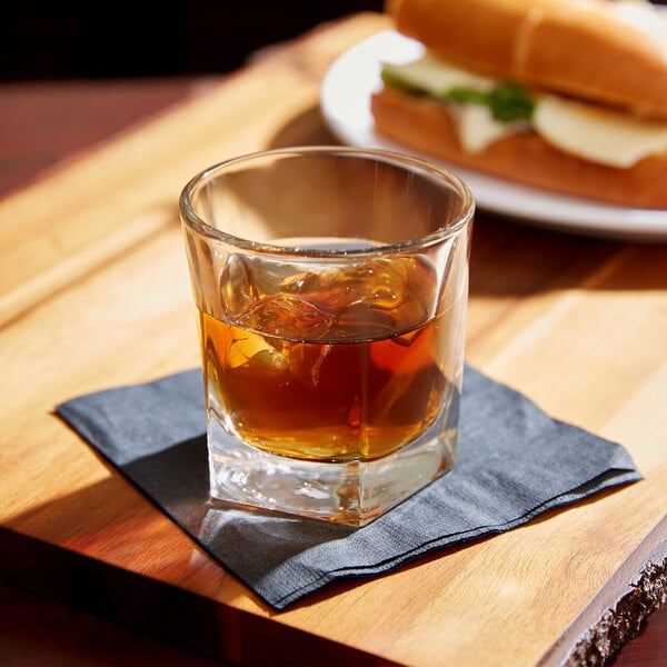 A Libbey Inverness rocks glass filled with brown liquid and ice on a table next to a sandwich.