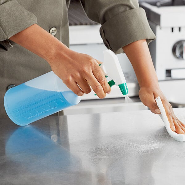 A person in a chef's coat using a white and green Lavex spray gun to clean a counter in a school kitchen.