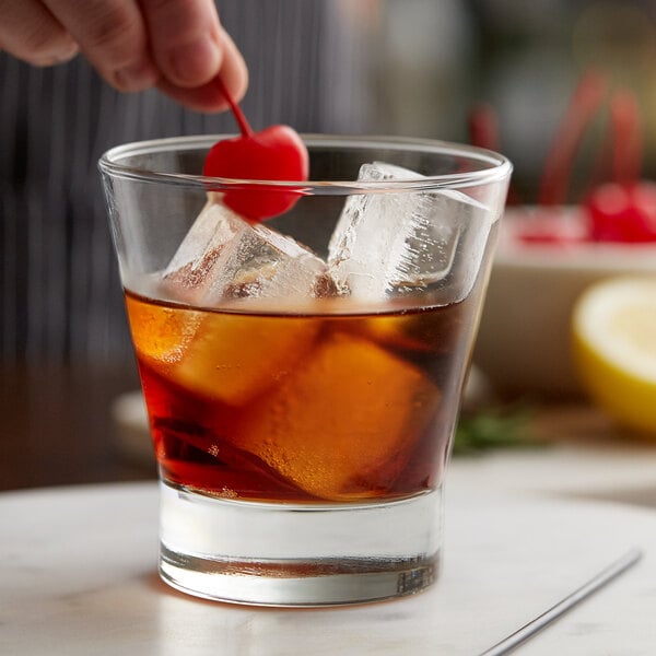 A hand holding a cherry over a Libbey old fashioned glass of ice with a cherry on top.