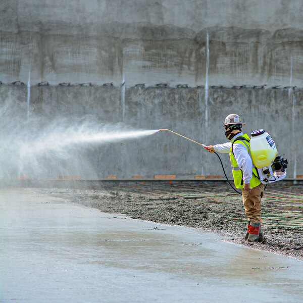 A man wearing a safety vest and using a Tomahawk backpack concrete sprayer to spray a concrete wall.