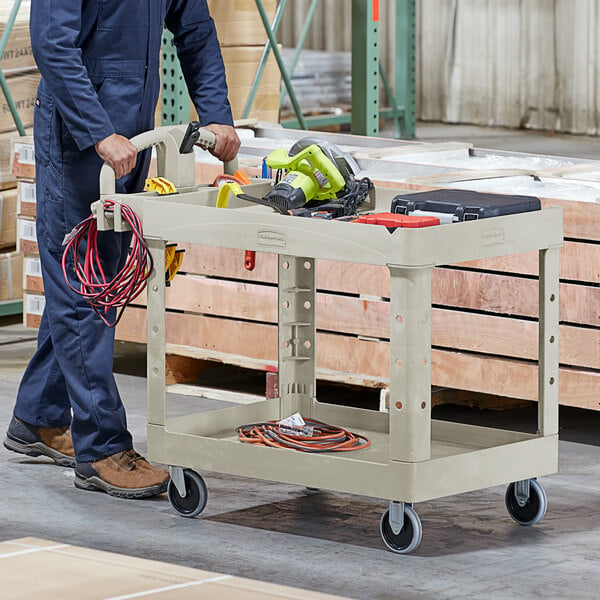 A man in a blue jumpsuit holding a Rubbermaid utility cart with a drill.