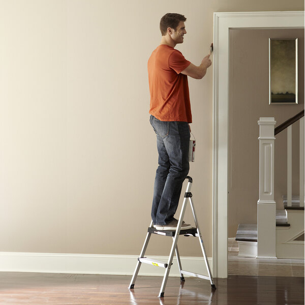 A man standing on a Cosco 2-step ladder holding a paint bucket.