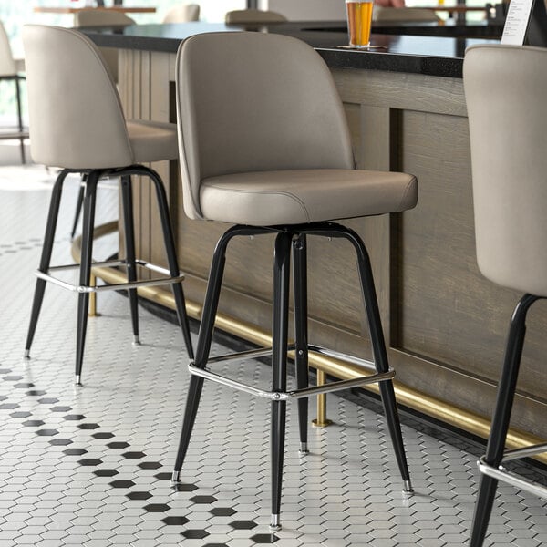 Three Lancaster Table & Seating gray vinyl bucket seat cushions on bar stools with black legs.