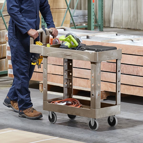 A man in blue overalls is pushing a Rubbermaid beige utility cart with tools on it.