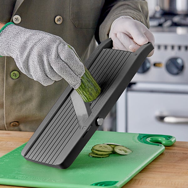 A gloved hand uses Emperor's Select large plastic mandoline to slice a cucumber.