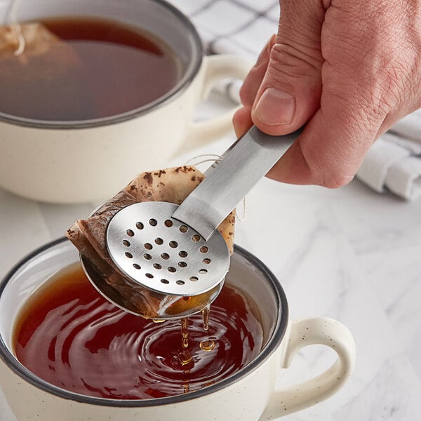 A person using Fox Run stainless steel tea bag tongs to hold a tea bag over a cup of tea.