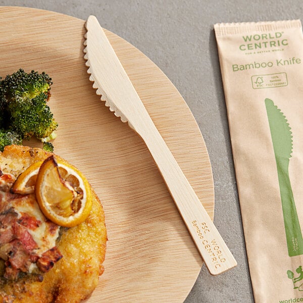 A wooden plate with a World Centric compostable bamboo knife on it next to a plate of food.