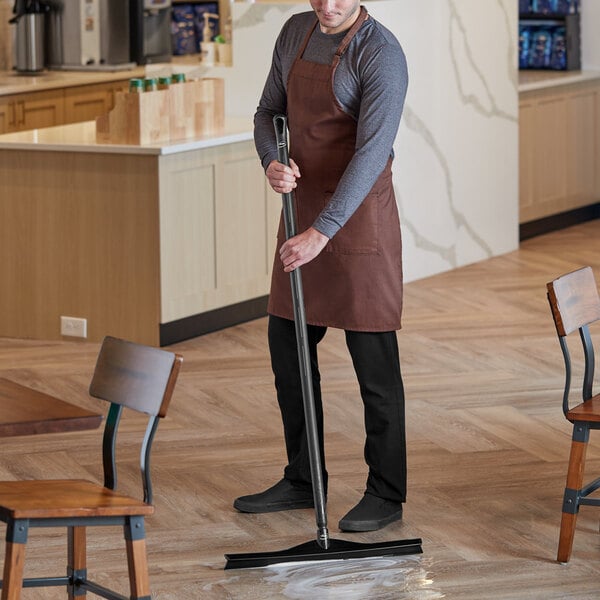 A man in an apron using a Vikan black single blade rubber floor squeegee with a long handle to clean a floor.
