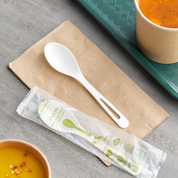 A wrapped World Centric compostable spoon on a table next to a cup of soup.