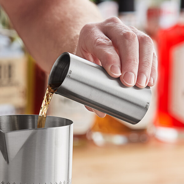 A person using an Outset stainless steel thimble jigger to pour liquid into a metal container.