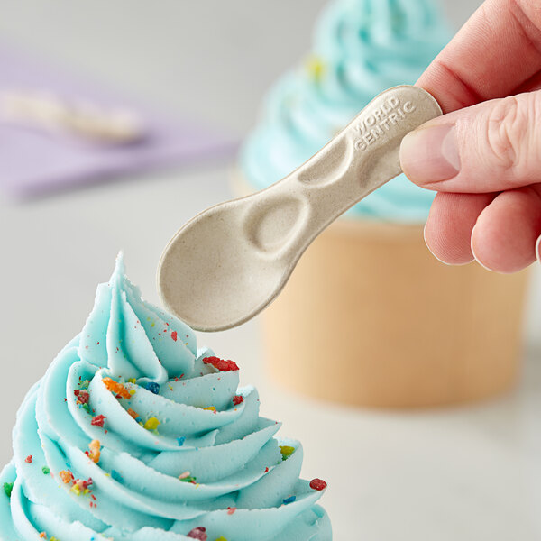 A hand holding a World Centric compostable fiber tasting spoon over a blue frosted cupcake.