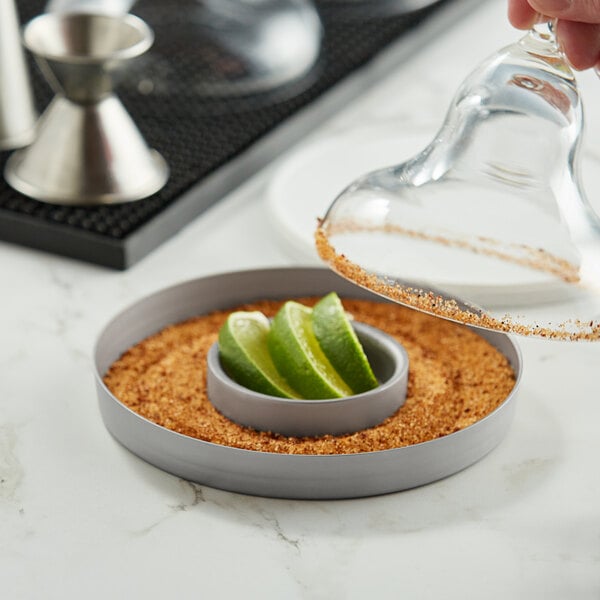 A person pouring lime juice into a circular dish of lime juice with a stainless steel Outset glass rimmer.