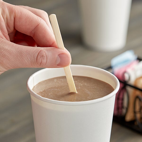 A hand holding a World Centric compostable bamboo stir stick in a cup of coffee.