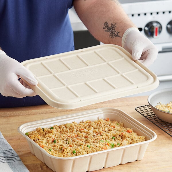 A person wearing gloves holds a white rectangular World Centric catering pan lid over a container of rice and vegetables.