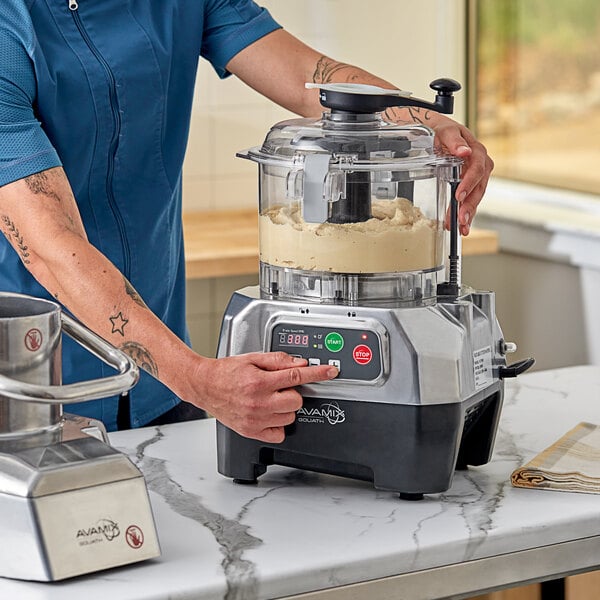 A commercial food processor with a clear plastic bowl and digital controls being operated on a kitchen counter.
