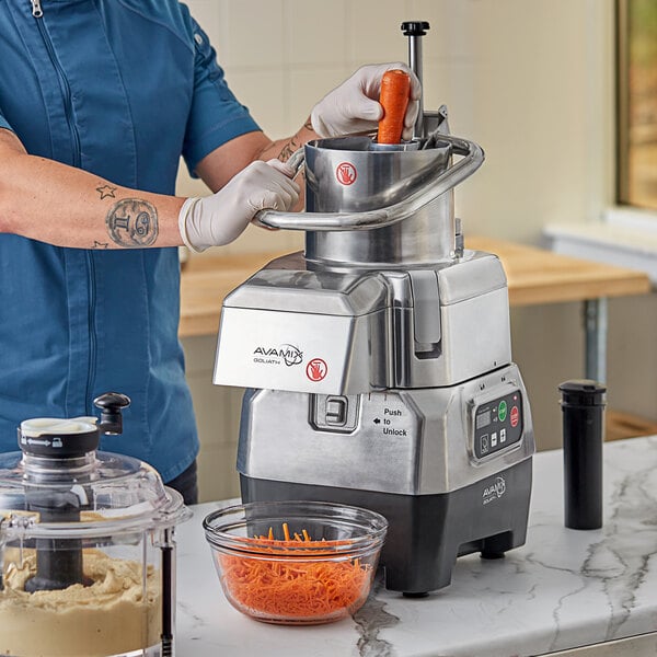A commercial combination food processor with a clear plastic bowl and continuous feed attachment, shown processing carrots.