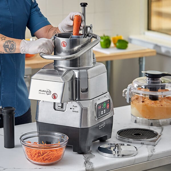 A commercial food processor with a stainless steel body, clear plastic bowl, and various attachments, shown in use for shredding carrots.