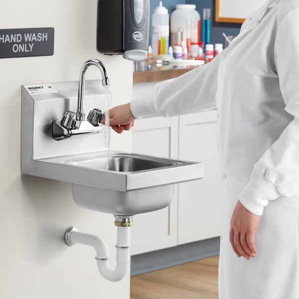 A woman in a lab coat washing her hands in a silver wall-mounted Regency hand sink over a counter.