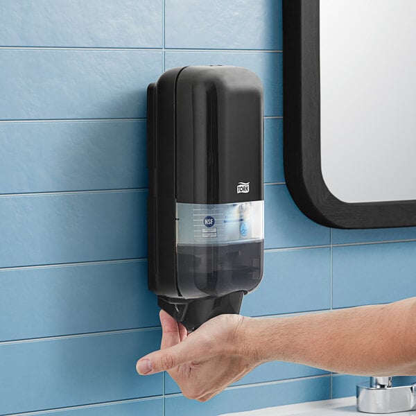 A person using a black Tork manual hand soap dispenser.