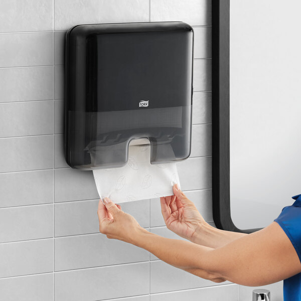 A woman using a Tork black plastic mini wall-mount multifold hand towel dispenser.