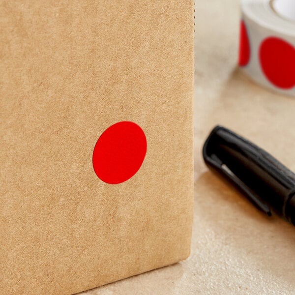 A brown box with a red circle on it next to a roll of Lavex Standard Red Semi-Gloss Paper Inventory Labels.