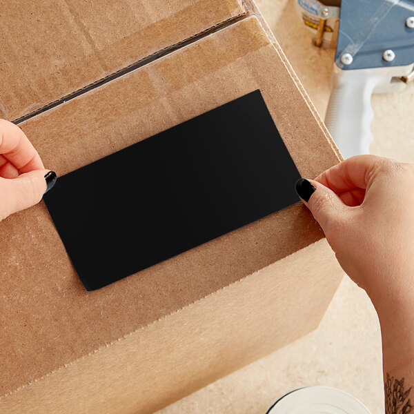 A person using a white and blue box cutter to open a cardboard box of black rectangular Lavex inventory labels.