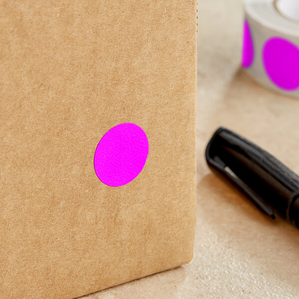 A close-up of a pen writing on a Lavex box with a fluorescent pink round label with purple writing.